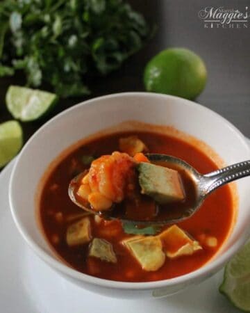 Caldo de Camarón, or Mexican Shrimp Soup in a white bowl with a rich, guajillo sauce. Spoon lifting shrimp and avocado. Cilantro and lime on a dark brown table.