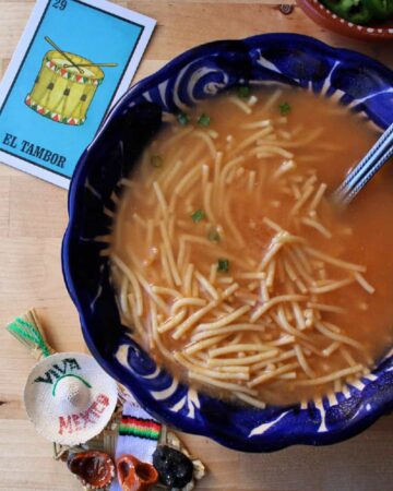 A blue bowl with Sopa de Fideo (Mexican Noodle Soup) surrounded by decorative Mexican toys.