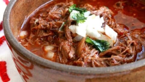 Birria de Res, or Mexican Beef Stew, in a brown bowl topped with chopped onions and cilantro.