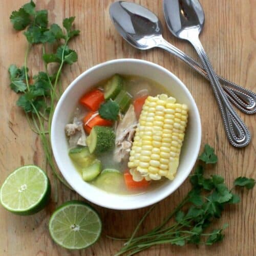 Caldo de Pollo, or Mexican Chicken Soup served in a white bowl and surrounded by lime and spoons.