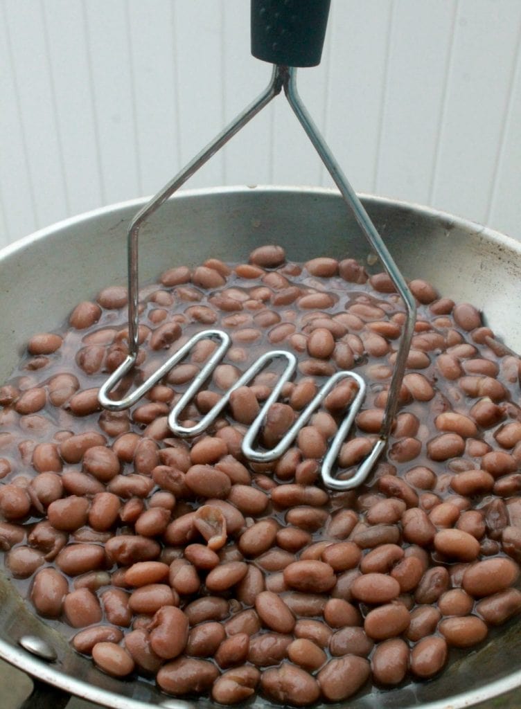 A potato masher mashing cooked beans in a metal skillet.