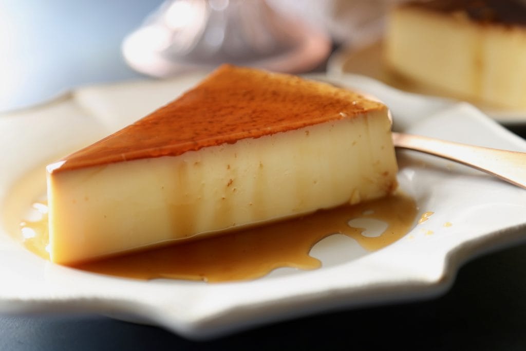 A slice of flan on a white plate drizzled with caramel sauce.