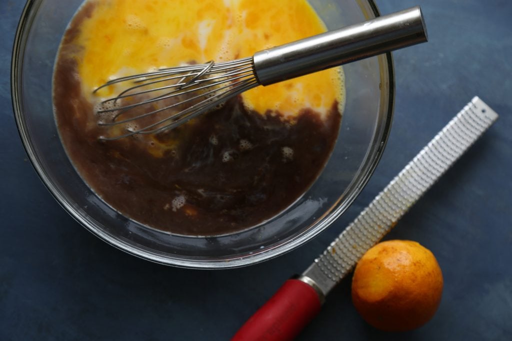 A glass bowl with a whisk next to a zester and an orange.