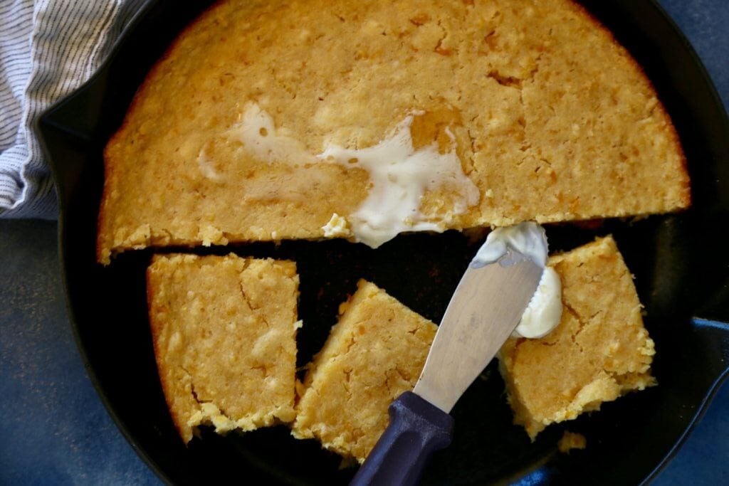 A skillet of golden pan de elote, partly sliced into squares. One square has butter melting on top, with a butter knife resting beside it. A striped cloth is visible at the edge of the skillet.