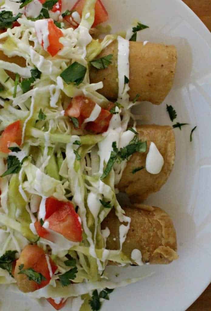 Potato and Cheese Taquitos Recipe topped with shredded cabbage, diced tomatoes, chopped cilantro, and Mexican crema
