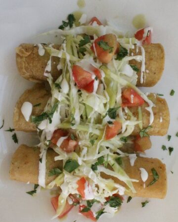 Taquitos on a white plate topped with shredded cabbage diced tomatoes chopped cilantro and drizzled with Mexican cream sauce