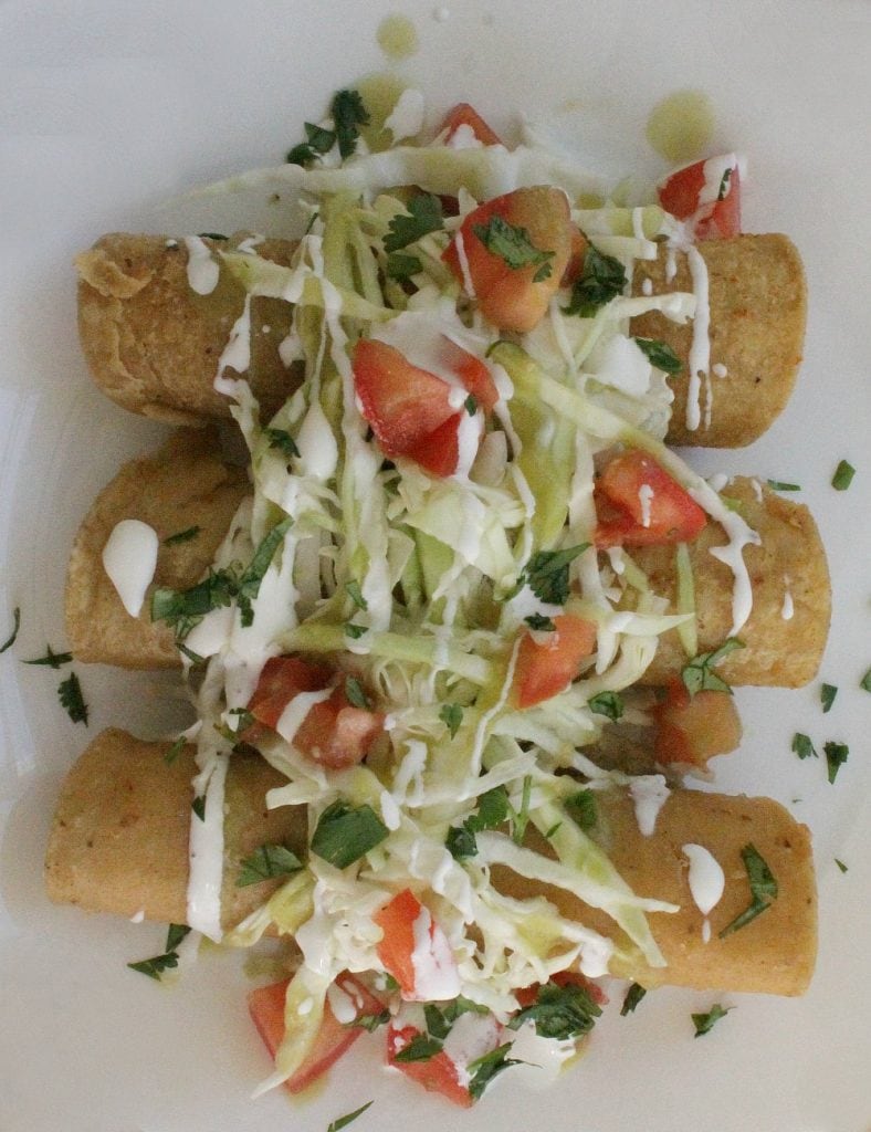 Taquitos on a white plate topped with shredded cabbage diced tomatoes chopped cilantro and drizzled with Mexican cream sauce