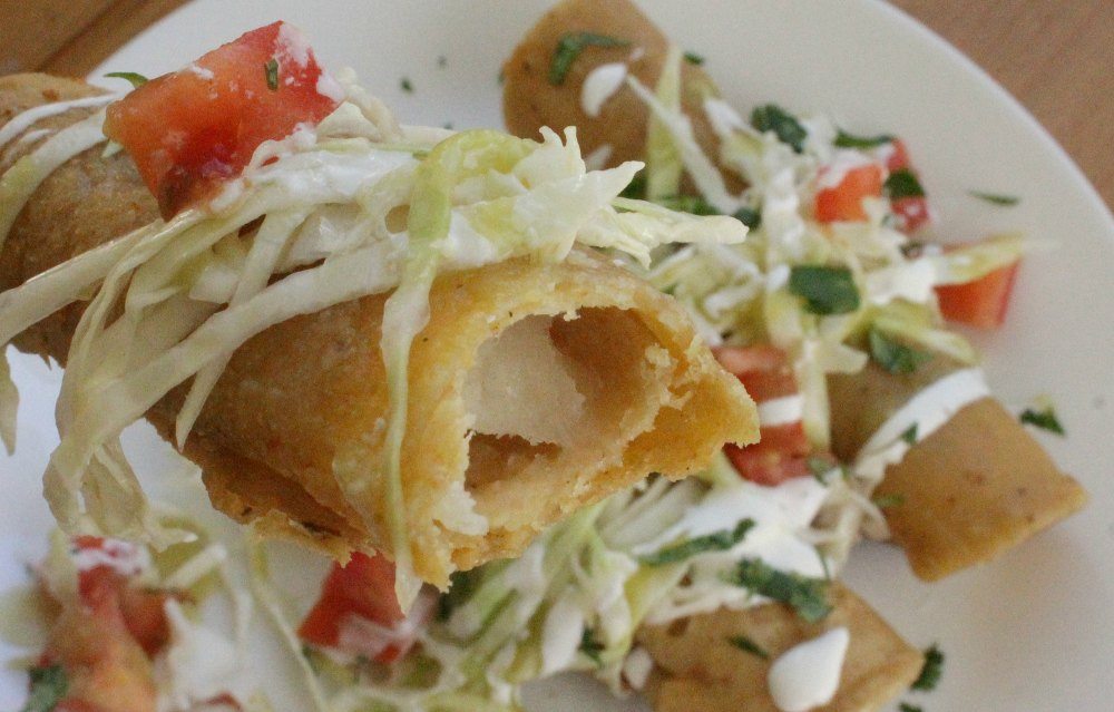 Close up picture, bite into the taquito, topped with cabbage and tomato. More taquitos on the plate in the distance