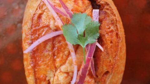 Torta Ahogada, or Drowned Sandwich, topped with pickled red onions and a cilantro leaf, surrounded by a spicy tomato sauce on a blue plate.