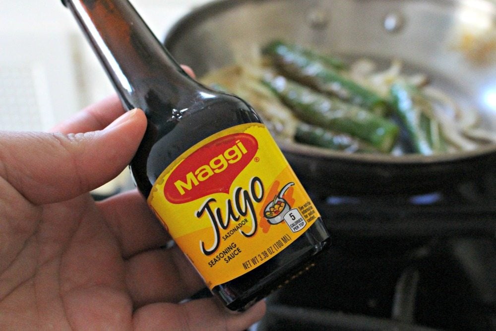 A hand holding a Jugo Maggi bottle with a skillet in the background.