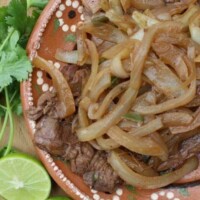 Bistec Encebollado (Mexican-Style Beef and Onions) served with lime wedges cilantro on decorative, clay plate