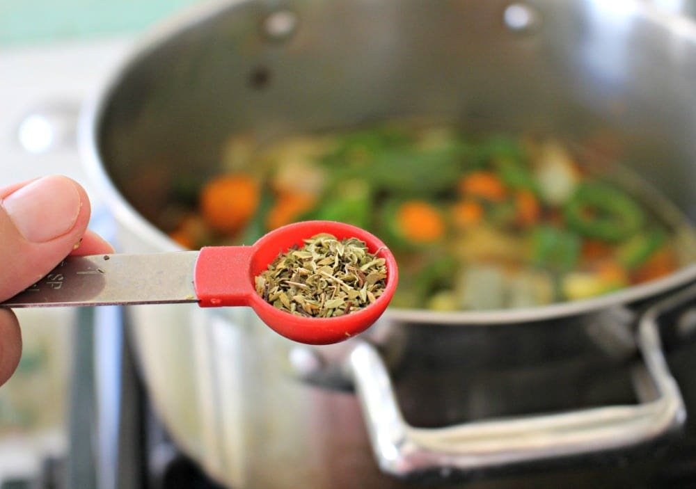 Measuring spoon with dried oregano over a stockpot with jalapenos.