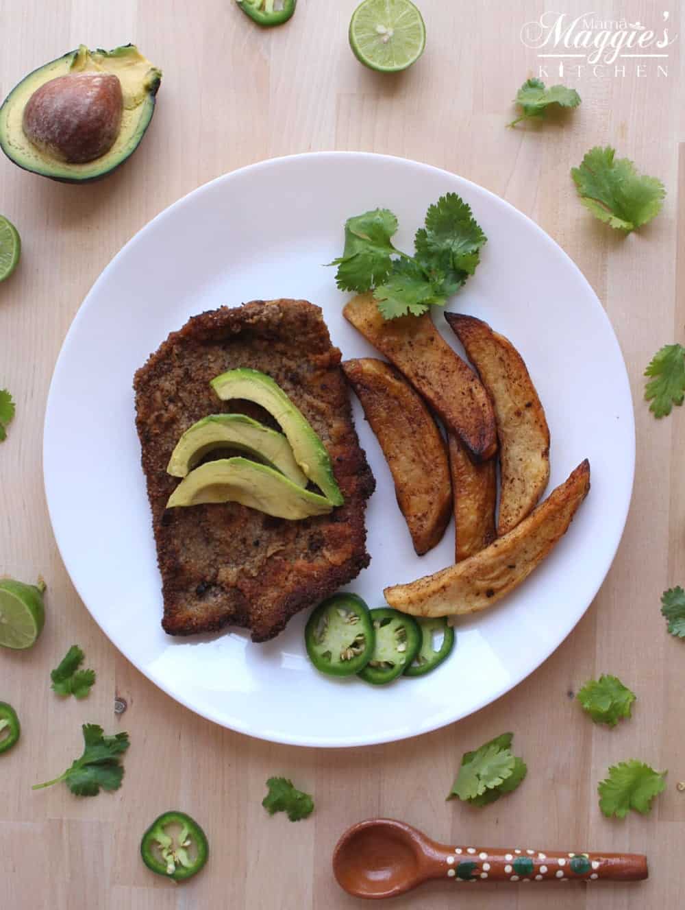 Milanesa de Res and homemade french fries sitting on a white plate. The meat is topped with avocado and surrounded by cilantro leaves.