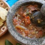 Roasted Tomato Salsa in a volcanic rock molcajete surrounded by jalapenos, chips, and a decorative Mexican cloth.