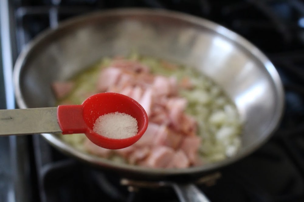 A red teaspoon of salt with the ham and onion skillet in the background. 