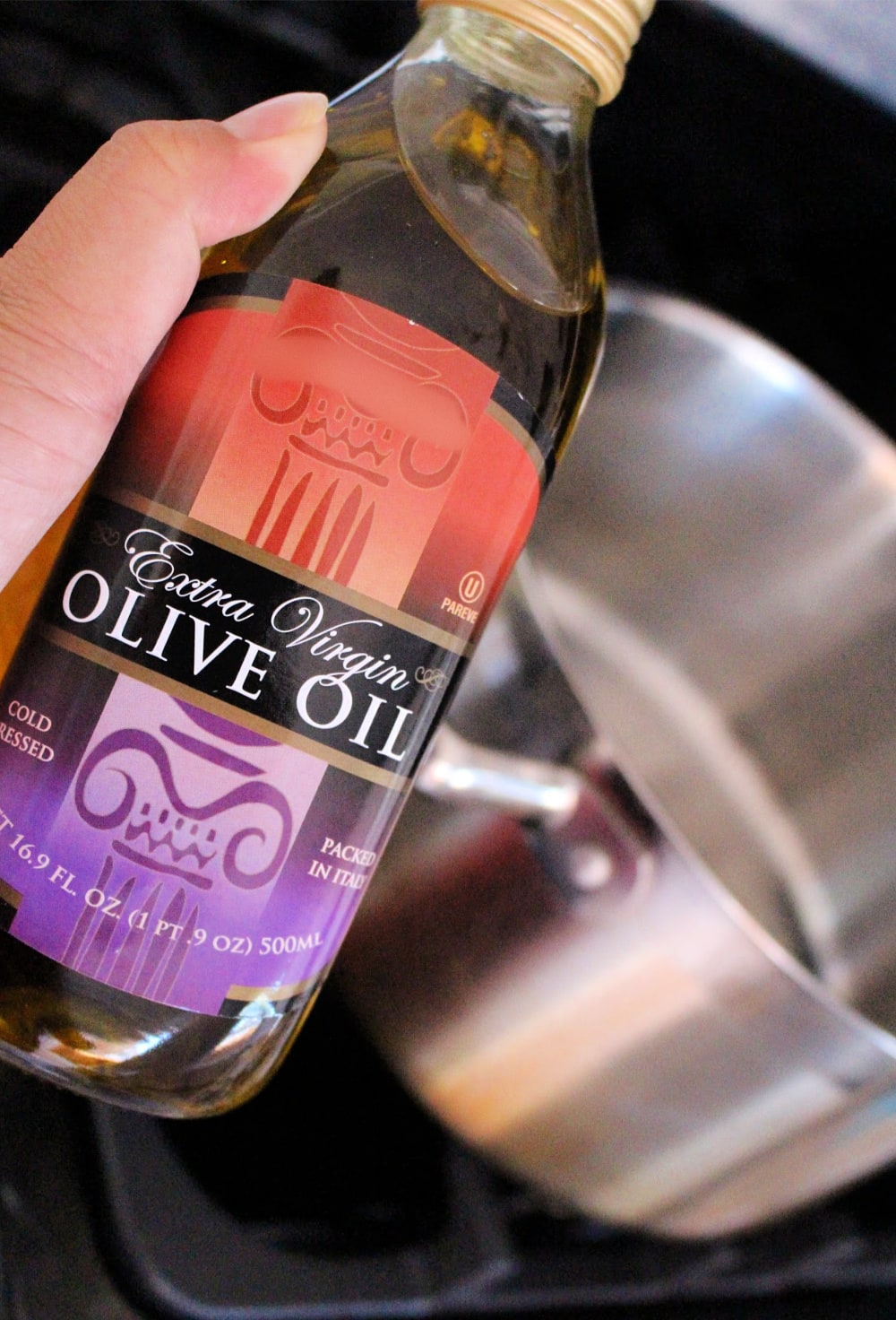 Hand holding a bottle of olive oil with a stock pot in the distance. 