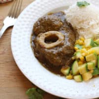 Chamorro en Salsa Verde, or Beef Hind Shank in Mexican green sauce, on a white plate with veggies and rice on a wooden surface next to a fork.