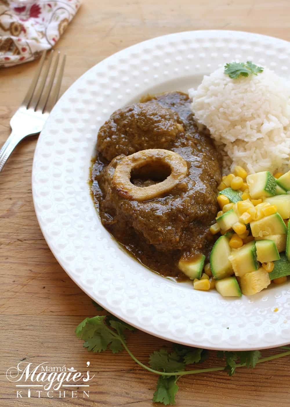 Chamorro en Salsa Verde, or Beef Hind Shank in Mexican green sauce, on a white plate with veggies and rice on a wooden surface next to a fork. 