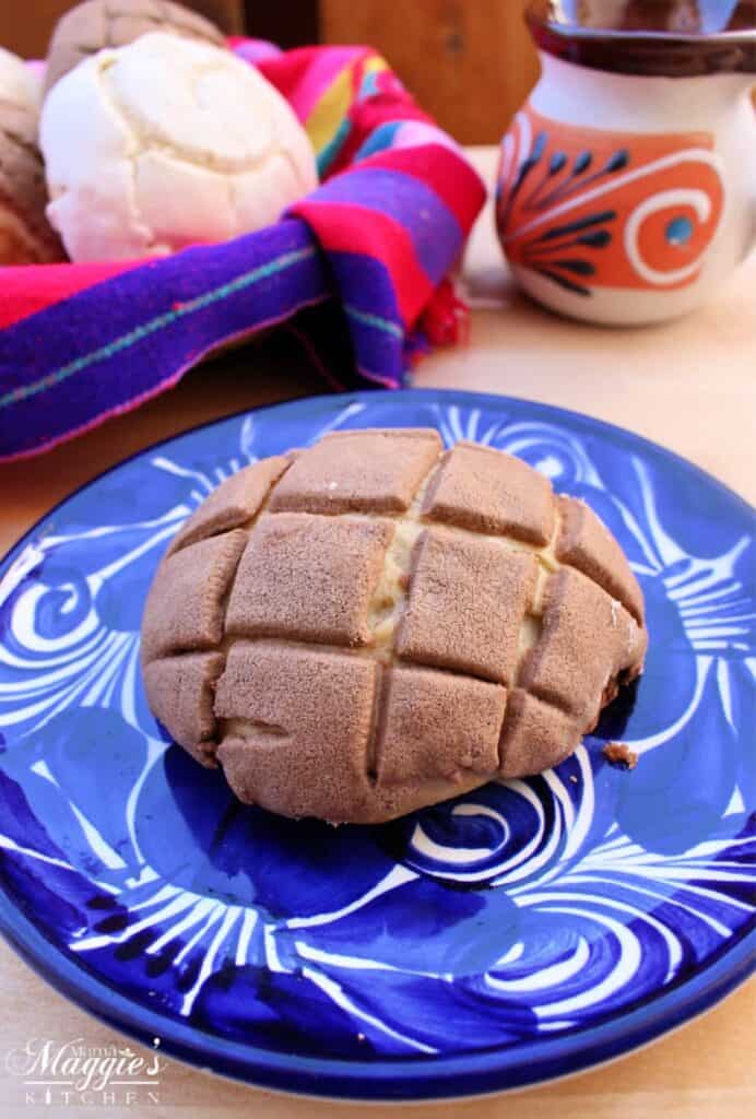Chocolate concha on a blue plate with a decorative Mexican basket in the background holding more conchas.