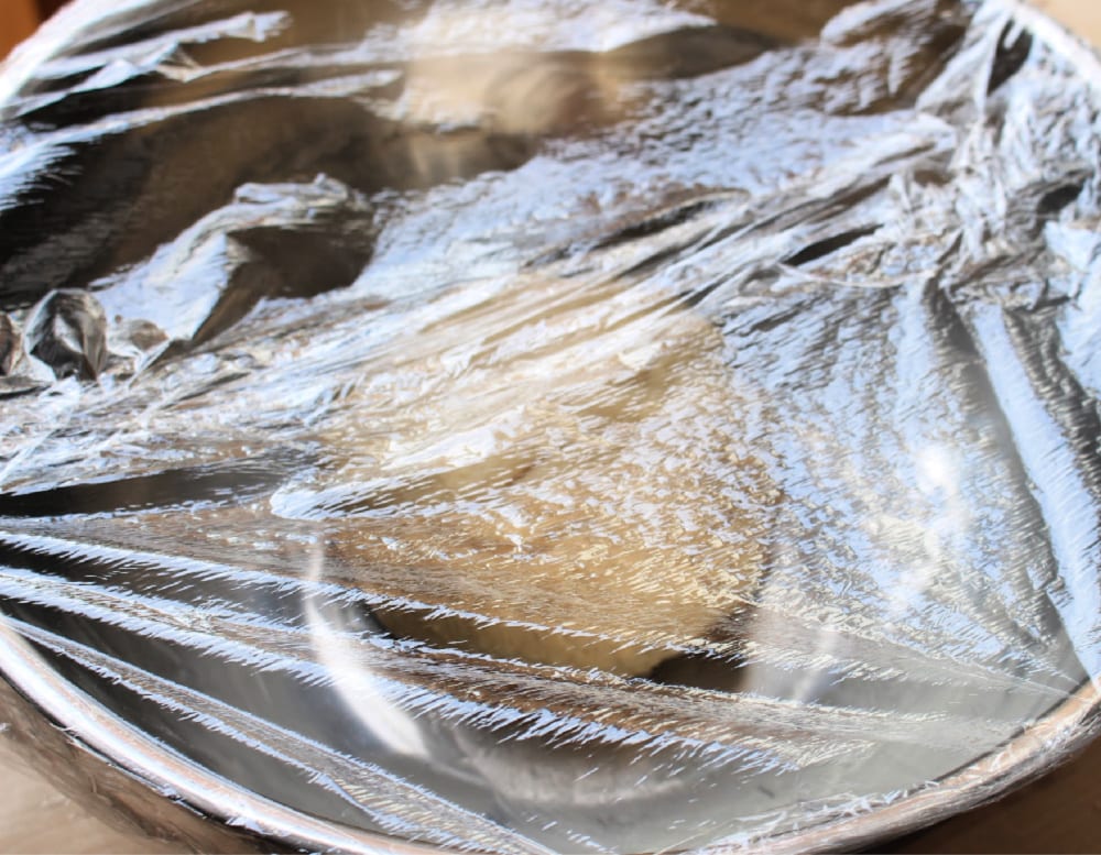 Dough in a metal bowl covered with plastic wrap. 