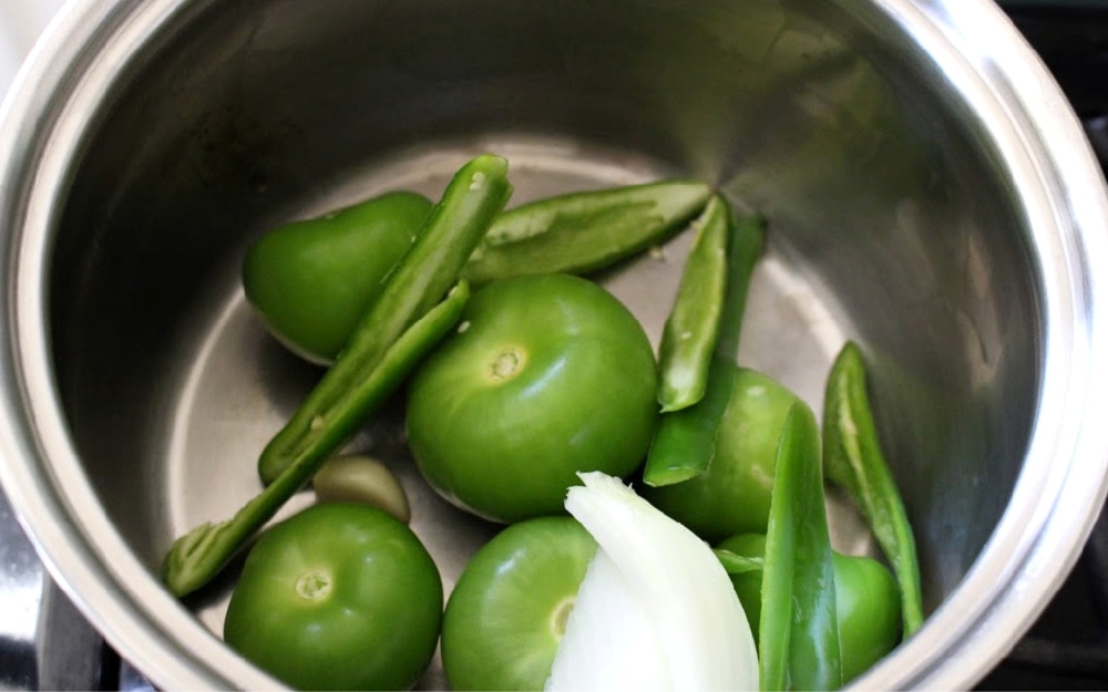 Green tomatillos, onion, and serrano peppers in a metal stock pot. 