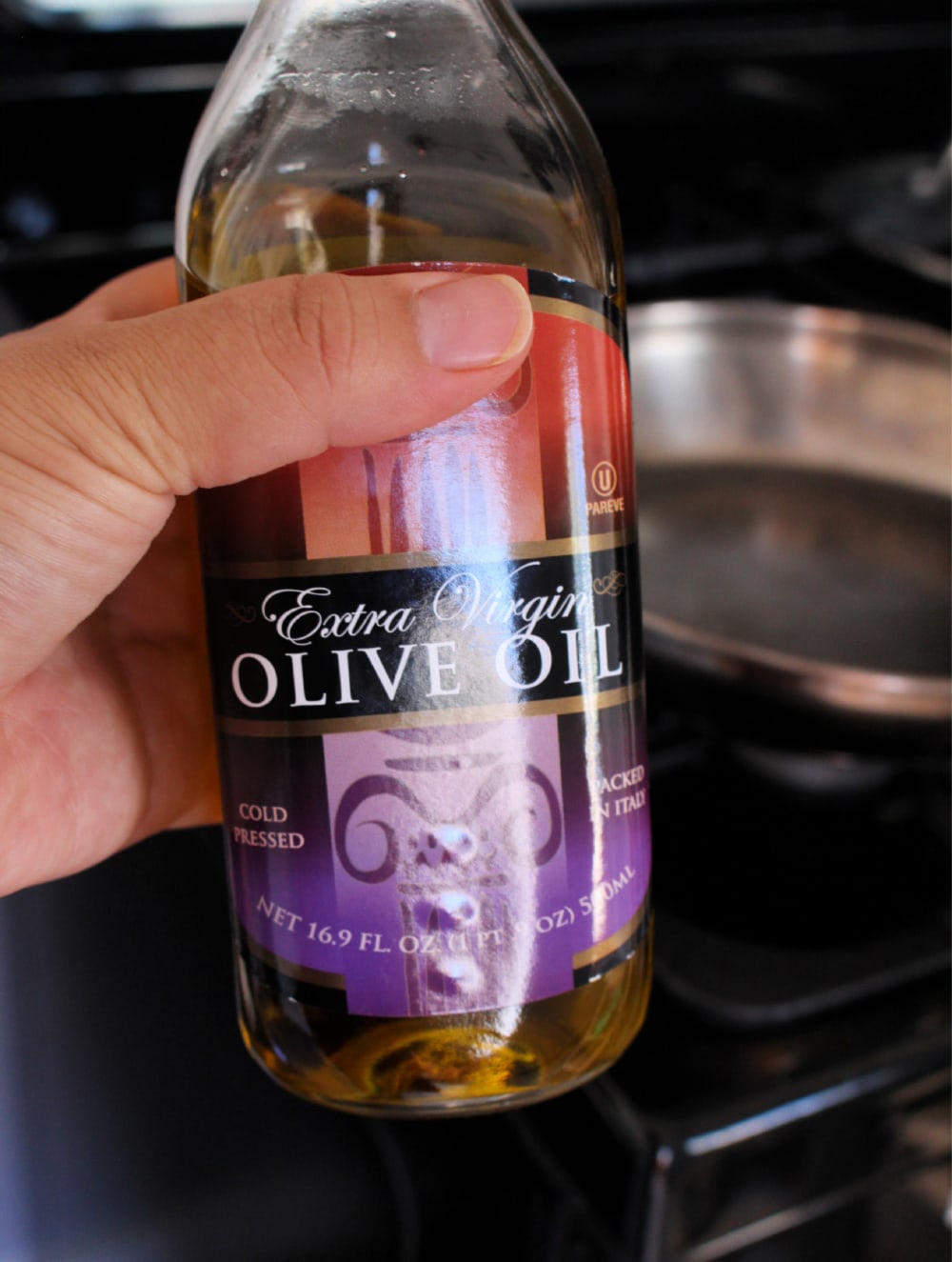 Hand holding a bottle of olive oil with a black stove in the background. 