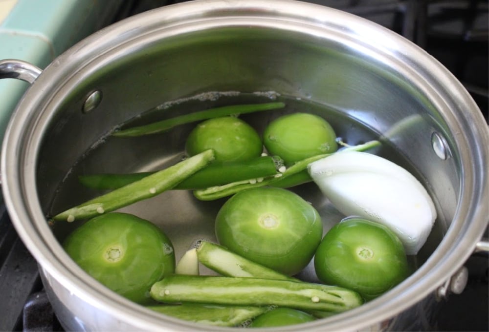 Tomatillos, serrano peppers, and onion floating in water in a stock pot.