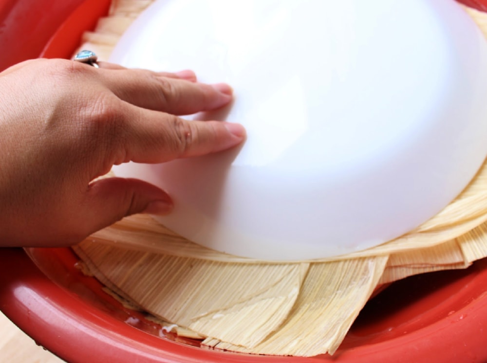 Hand over white plate submerging the corn husks in a large red container