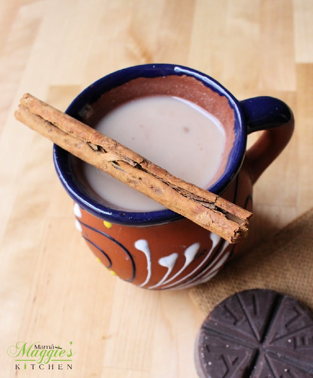 Champurrado in a decorative Mexican clay cup topped with a cinnamon stick next to a tablet of Mexican chocolate.