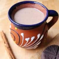 Champurrado in a decorative, clay Mexican cup surrounded by a cinnamon stick and Mexican chocolate tablet.