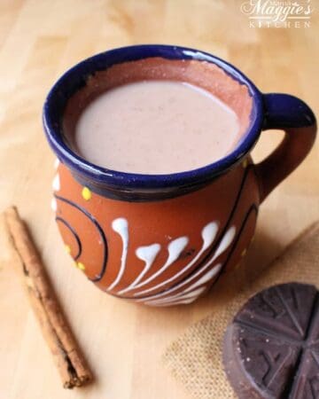 Champurrado in a decorative, clay Mexican cup surrounded by a cinnamon stick and Mexican chocolate tablet.