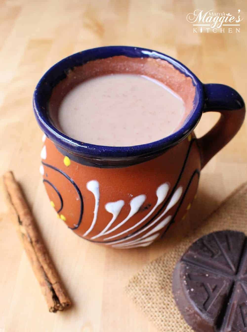 Mexican Champurrado in a decorative, clay cup surrounded by a cinnamon stick and Mexican chocolate tablet.