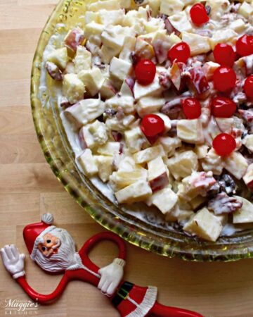 Ensalada Navideña (or Mexican Christmas Fruit Salad) in a green glass bowl topped with cherries and next to a toy Santa doll.