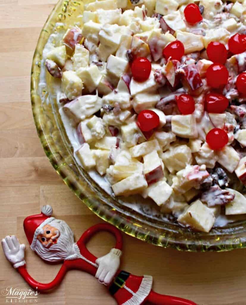 Ensalada Navideña (or Mexican Christmas Fruit Salad) is a mixture of apples and other fruits covered in a dreamy and creamy dressing in a green, glass bowl next to a plastic Santa.