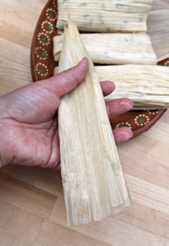 Hand folding corn husks with a pile of already assembled tamales to the side.