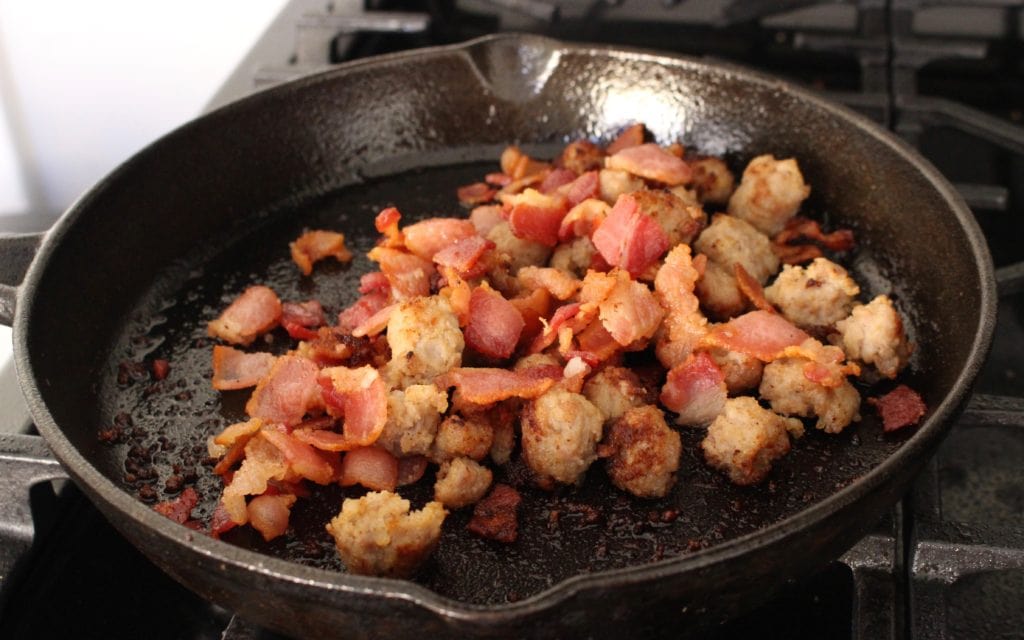 Sausage and Bacon drained and cooking in a black iron skillet.