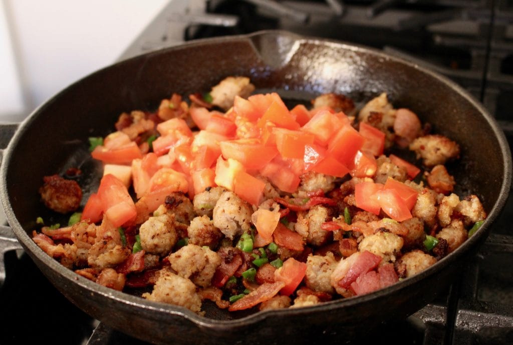 Diced tomato, bacon, and sausage all cooking together in a skillet.