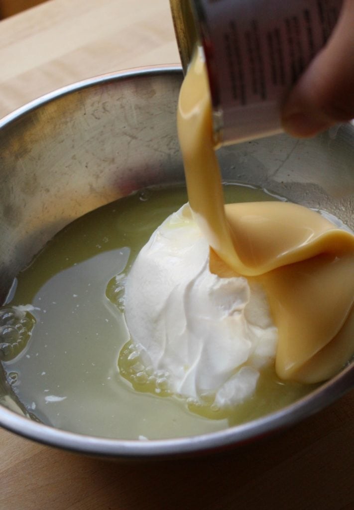 Sweetened condensed milk pouring into a metal bowl with sour cream and pineapple juice.