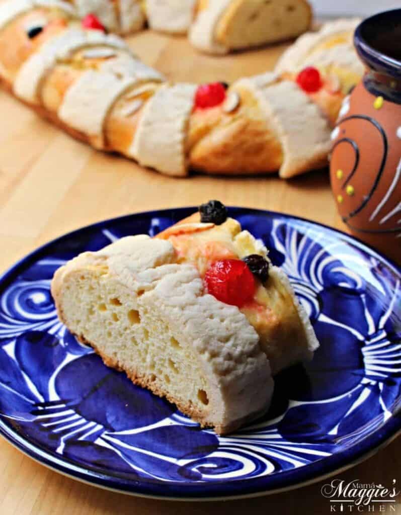 A slice of Rosca de Reyes (or Roscón de Reyes) on a decorative blue plate with the cake in the background.