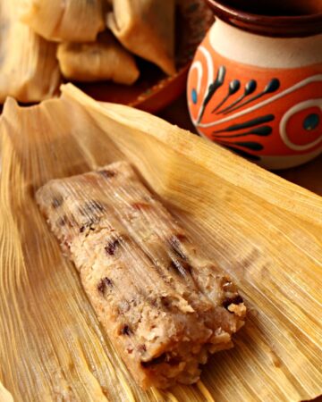 Tamales Dulces (Sweet Tamales) unwrapped but still in the corn husk surrounded by a pile of tamales and a decorative Mexican cup.