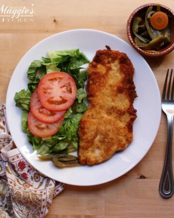 Milanesa de Pollo (Chicken Milanese) on a white plate next to a salad and surrounded by a decorative napkin and jalapeno slices and fork.