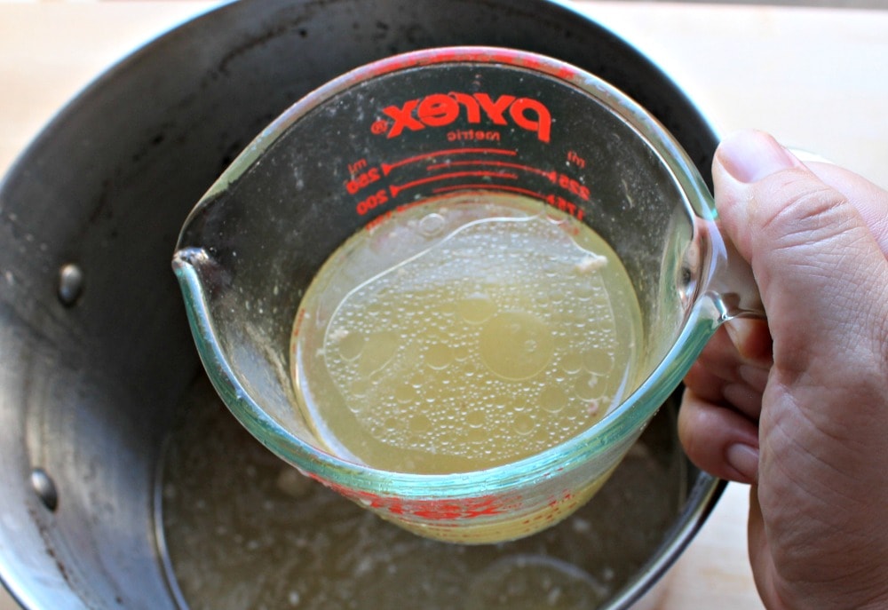 Hand holding a measuring cup with pork broth over the stock pot used to cook the pork for the tamales.