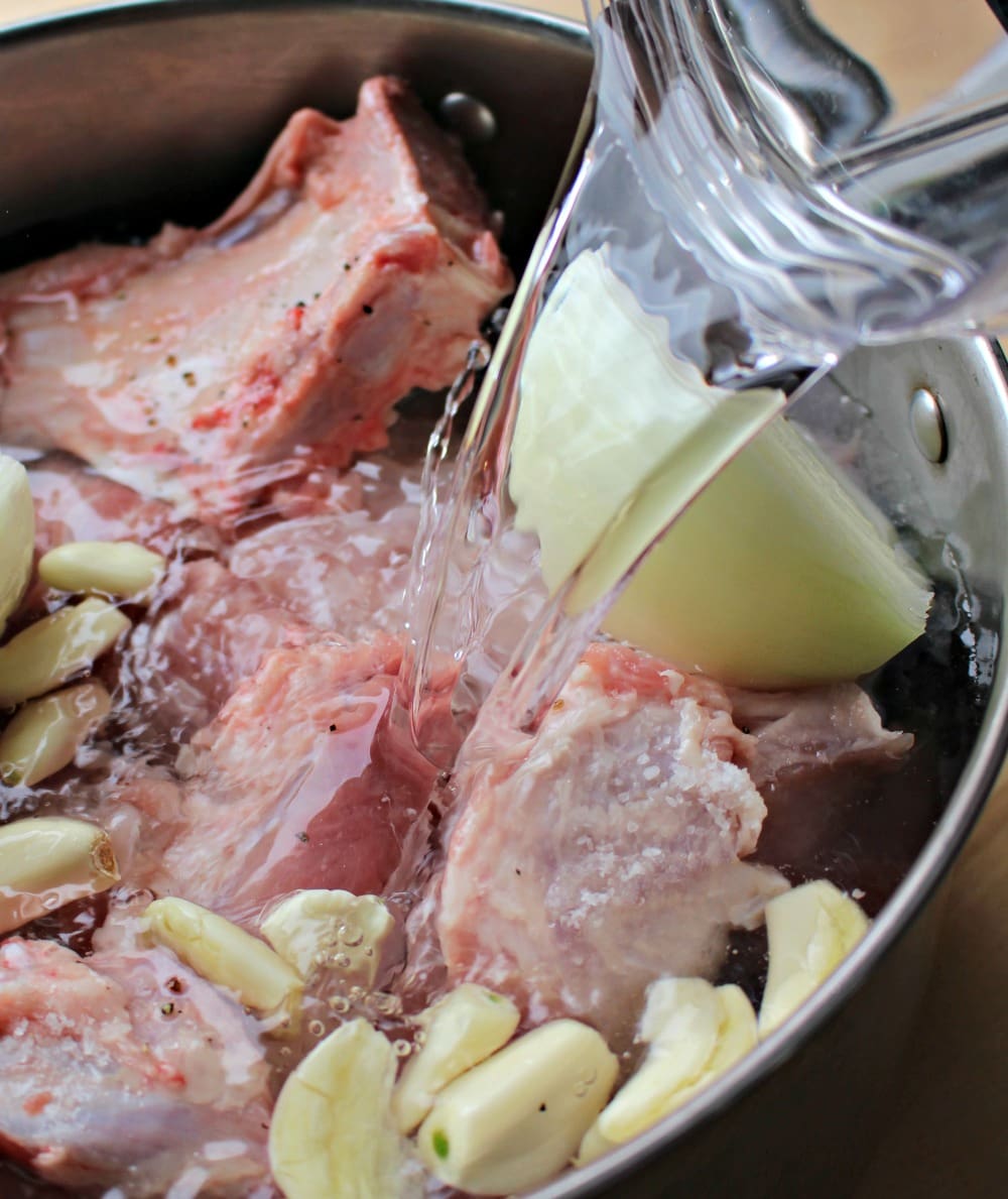 Water pouring inside a pot of raw pork pieces and lots of garlic cloves.