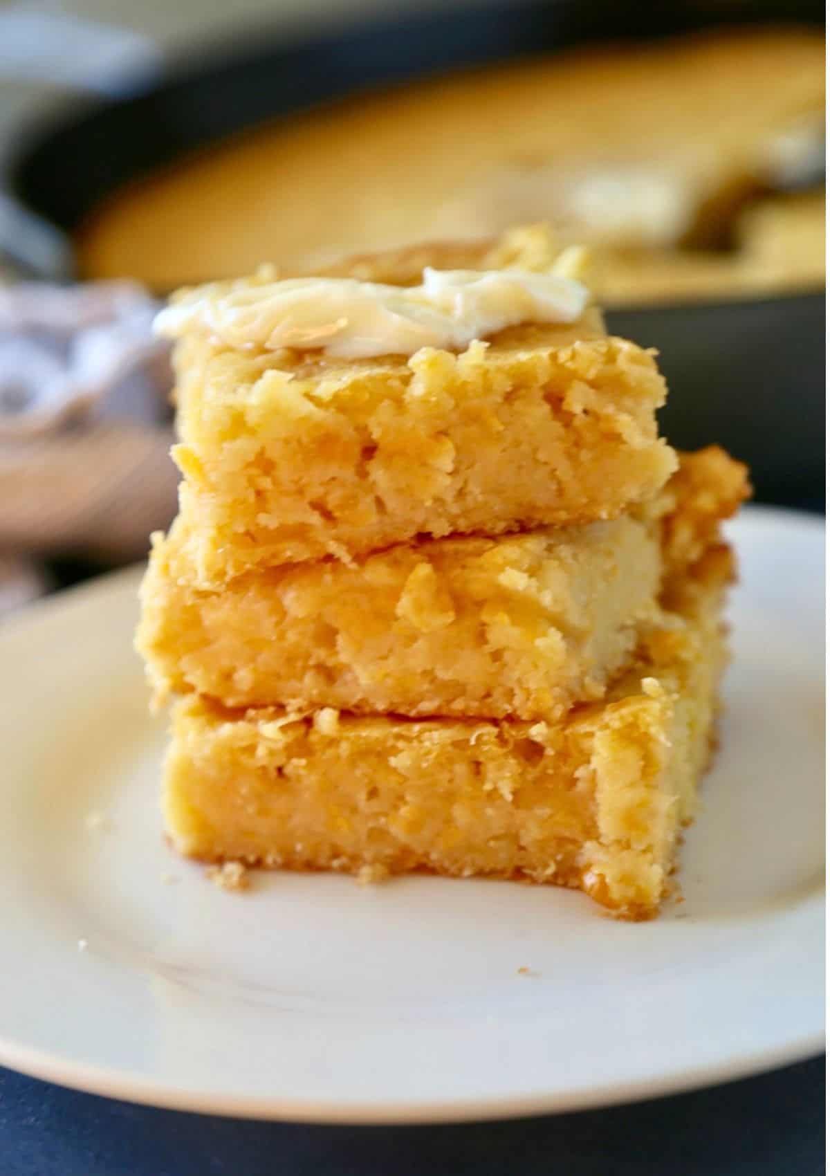 A stack of three golden, crumbly cornbread squares, reminiscent of pan de elote, sits on a white plate with a pat of melted butter on top. In the background, a pan of more cornbread is softly blurred.