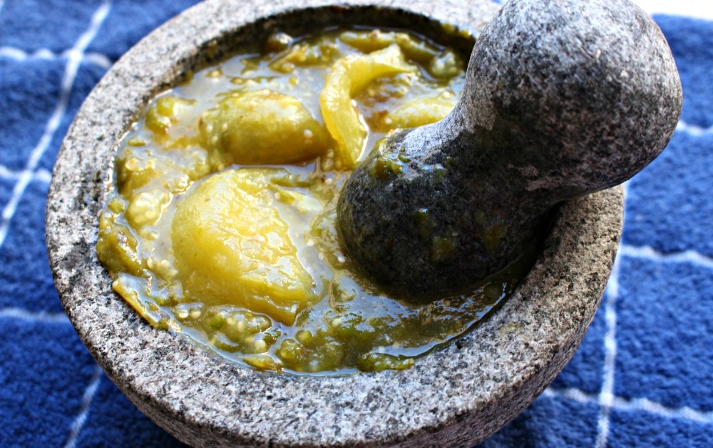 Tomatillos being smashed in a molcajete sitting on a blue towel.
