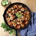 Camarones al Ajillo (Mexican Shrimp in a spicy garlic sauce) in a black iron skillet topped with lime slices and surrounded by green cilantro leaves.