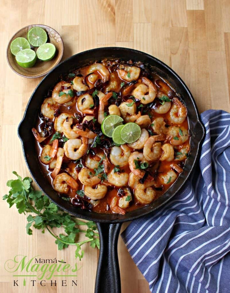 Camarones al Ajillo (Mexican Shrimp in a spicy garlic sauce) in a black iron skillet topped with lime slices and surrounded by green cilantro leaves.