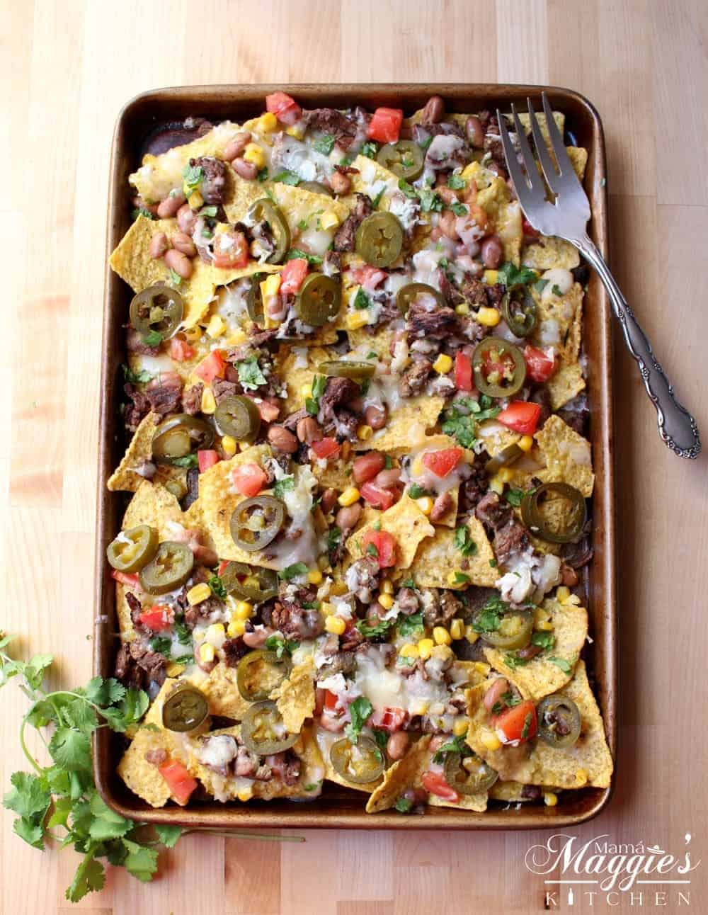 Carne asada nachos on a sheet pan next to a serving fork and cilantro.