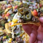 Hand holding a tortilla chip with pickled jalapenos, carne asada, and melted cheese over the rest of the carne asada nachos.