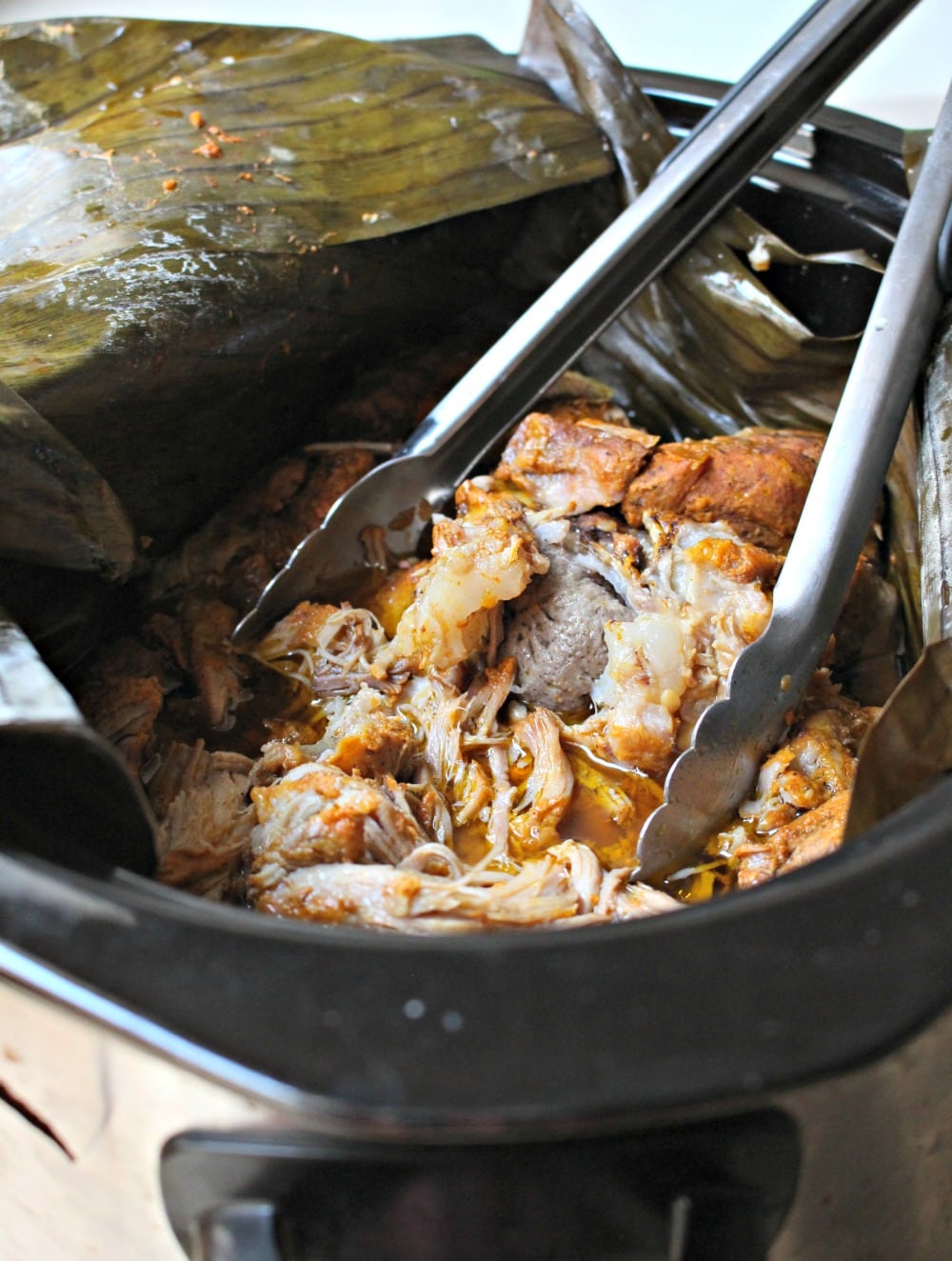 Cooked cochinita pibil in a crock pot with the banana leaves opened and tongs inside.